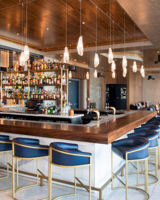 A modern bar with blue bar stools, hanging lights, a stocked liquor shelf, and additional seating areas in a stylish, contemporary setting.
