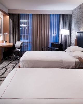 A modern hotel room with two neatly made beds, a large flat-screen TV, a desk with a chair, and stylish decor featuring a patterned wall.