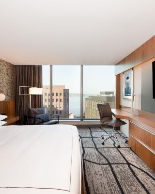 A modern hotel room with a large bed, wall-mounted TV, a desk and chair, patterned wallpaper, and a window view of buildings outside.