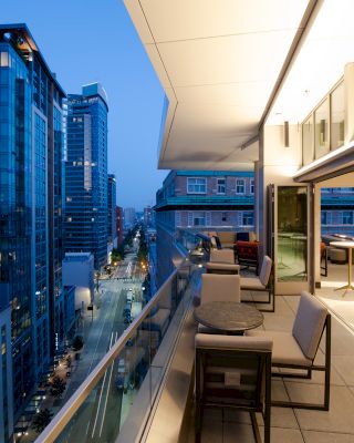The image shows a modern cityscape view from a balcony at dusk, with tall buildings and a sleek, stylish outdoor seating area with tables and chairs.