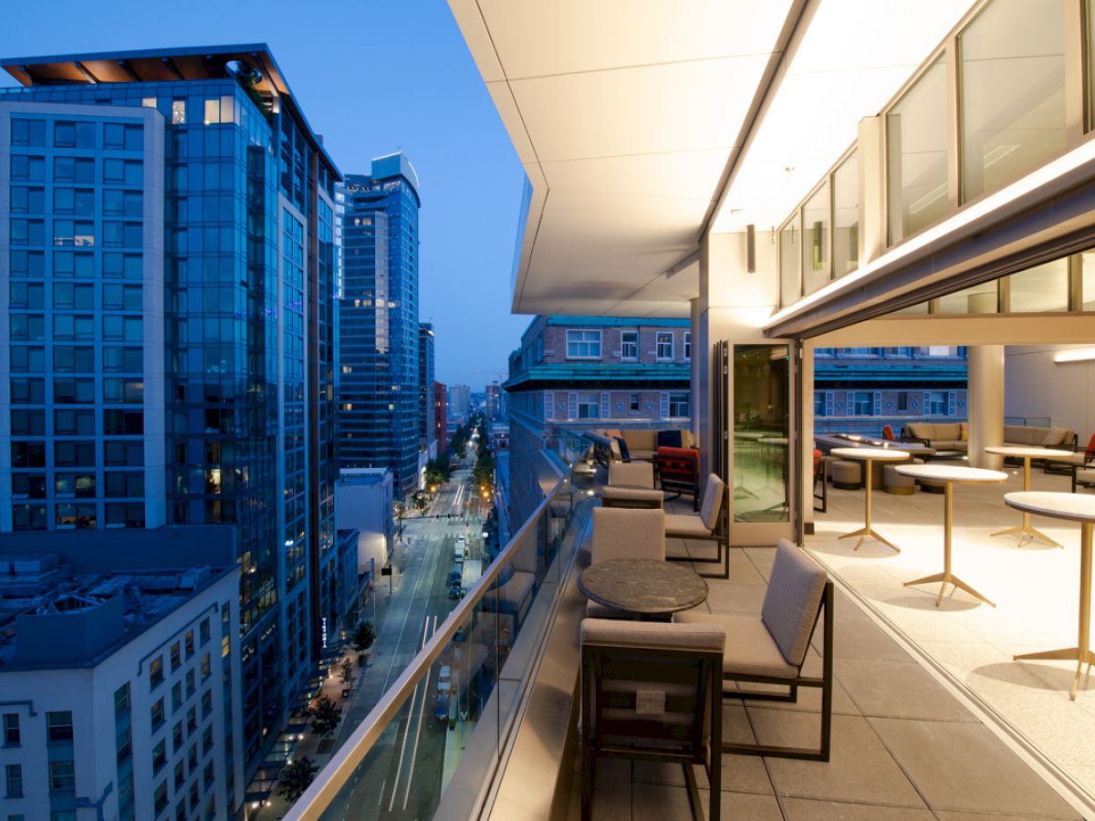 This image shows a modern balcony with chairs and tables, overlooking a cityscape with high-rise buildings and a street below, at dusk.