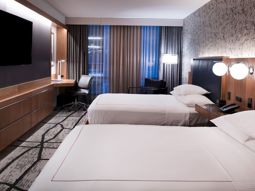 This image shows a modern hotel room with two double beds, a wall-mounted TV, a work desk with a chair, and large windows with curtains ending the sentence.