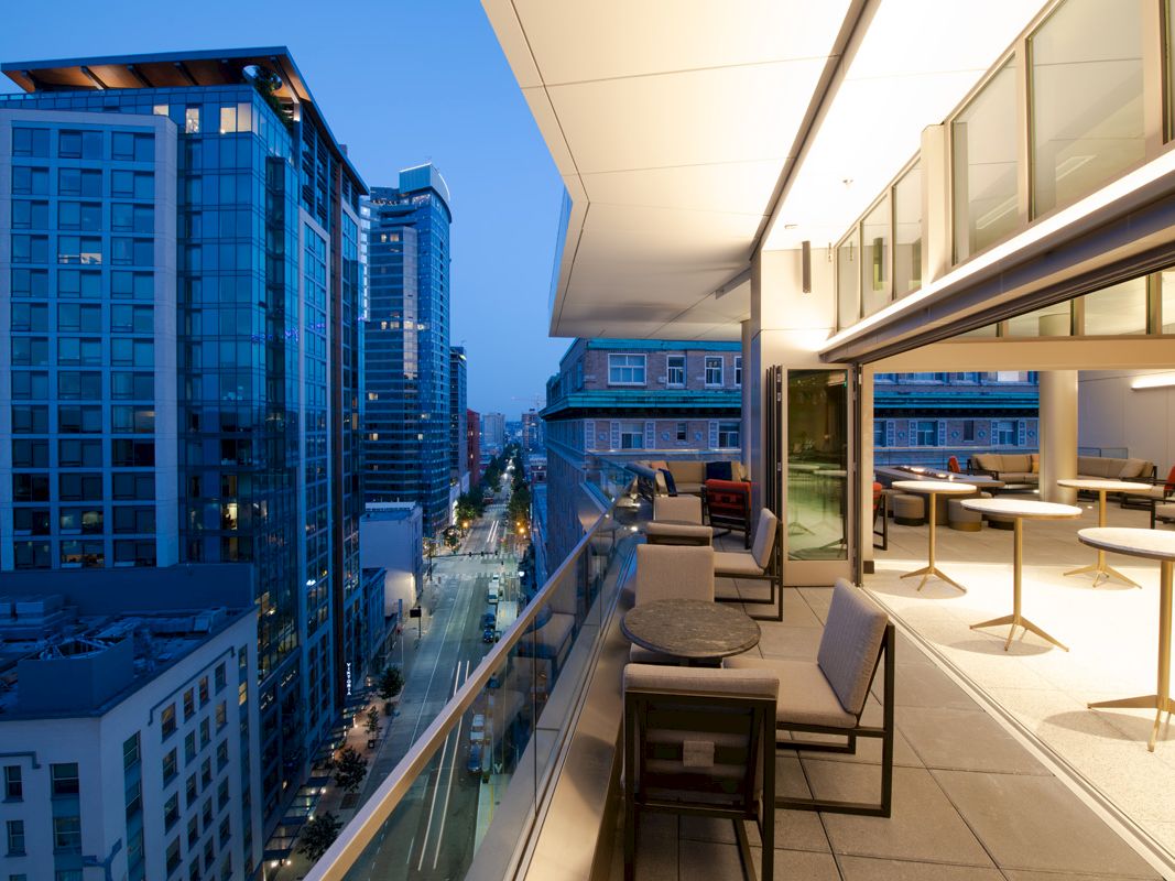 This image shows a balcony view of a modern cityscape at dusk, with high-rise buildings, a street below, and an outdoor seating area with tables and chairs.