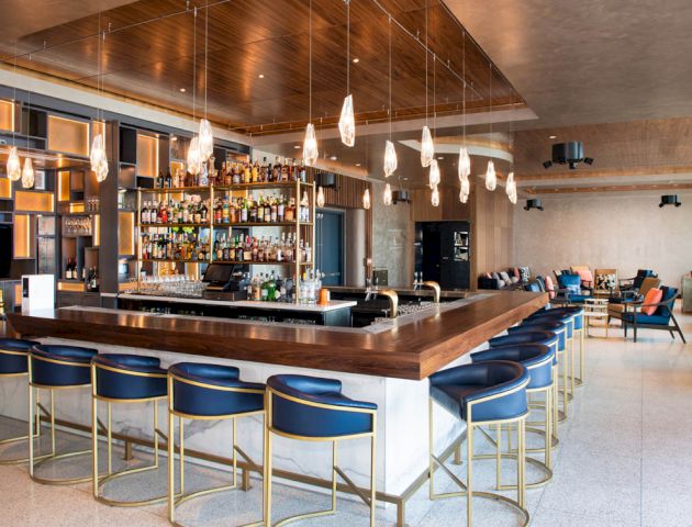 The image shows a modern bar with a central counter, blue upholstered chairs, hanging lights, and shelves stocked with bottles.