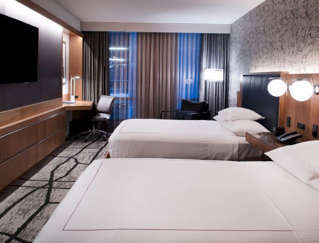 A modern hotel room with two double beds, a large TV, a work desk, and contemporary decor and lighting, creating a comfortable and stylish atmosphere.