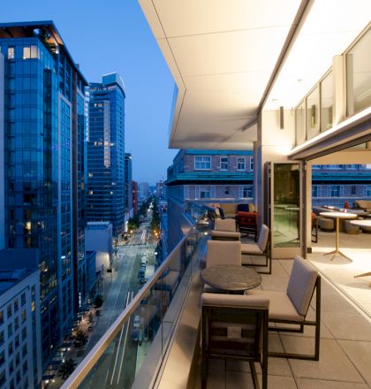 A modern rooftop terrace with outdoor seating overlooks a city street lined with tall buildings, illuminated as evening approaches.
