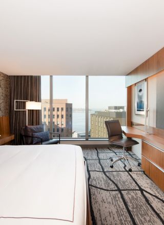 A modern hotel room features a large bed, a desk with a chair, a wall-mounted TV, and a window with a city view.