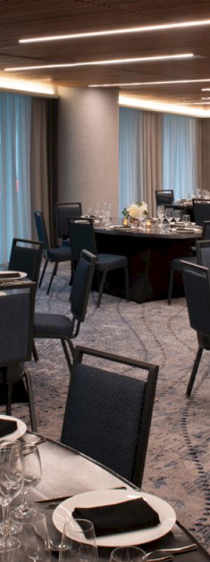 The image shows an elegant dining area with round tables setup for an event, featuring black chairs, table settings with wine glasses, and white floral centerpieces.