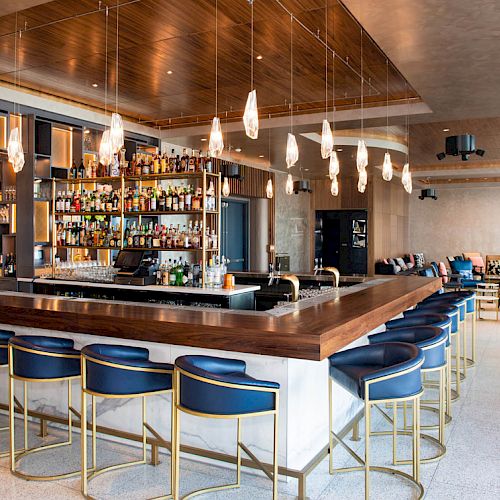 A modern bar with blue and gold stools, a wide selection of bottles, pendant lighting, and a seating area in the background.