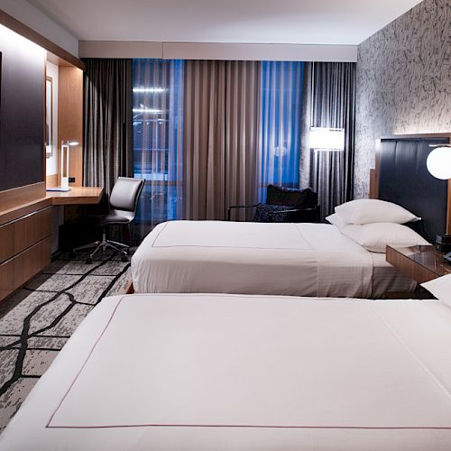 A modern hotel room with two neatly made beds, a wall-mounted TV, a desk, chairs, and well-designed lighting.