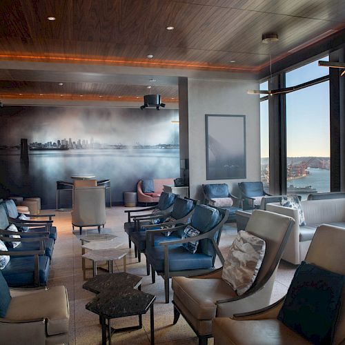 The image shows a modern lounge with comfortable seating, large windows with a waterfront view, and a wall mural depicting a city skyline.