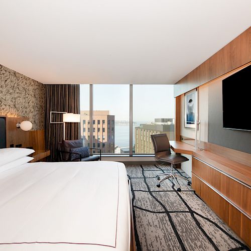 A modern hotel room with a large bed, TV, desk, chair, and a city view through floor-to-ceiling windows, featuring contemporary decor and furnishings.