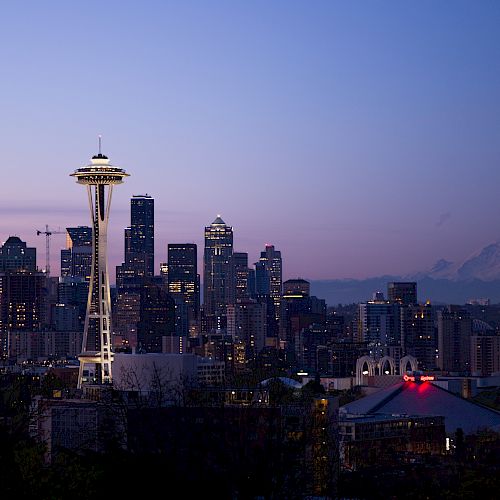A city skyline at twilight with the Space Needle prominently visible, set against a backdrop of mountains and illuminated buildings.