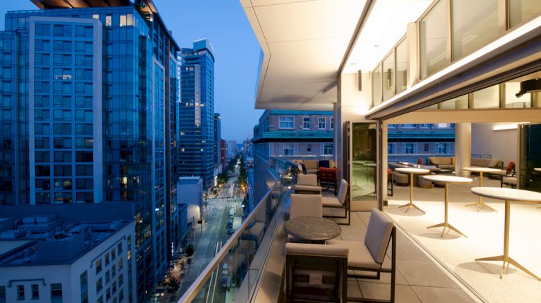 This image shows a modern cityscape with tall glass buildings. A well-furnished balcony overlooking the street features tables and chairs.