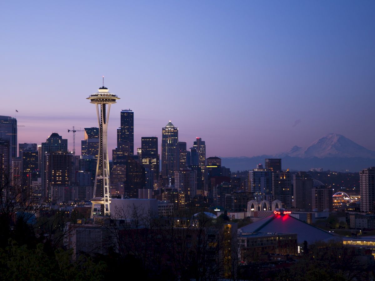 The image shows the Seattle skyline at dusk, featuring the Space Needle and Mount Rainier in the background, under a clear sky.