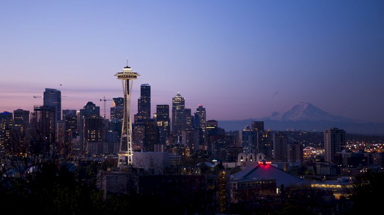 The image shows the Seattle skyline at dusk, featuring the Space Needle and Mount Rainier in the background, under a clear sky.