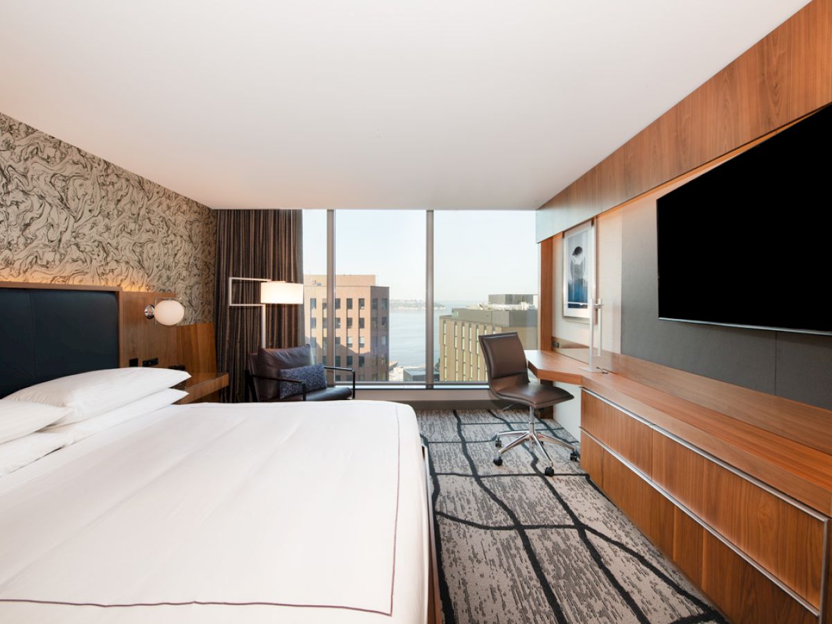 A modern hotel room with a large bed, desk, flat-screen TV, and large window offering a city view. The room has contemporary decor and furnishings.