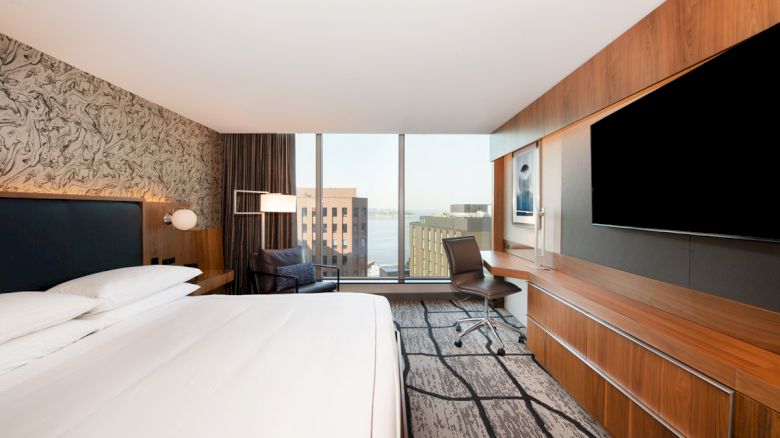 A modern hotel room with a large bed, desk, flat-screen TV, and large window offering a city view. The room has contemporary decor and furnishings.