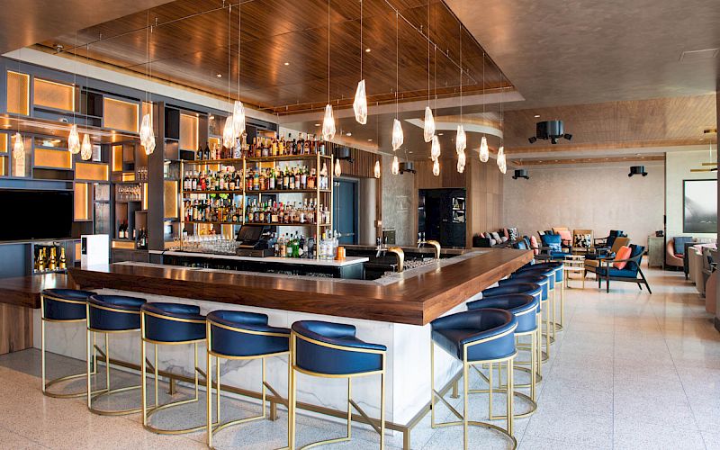 This image shows a modern bar with blue stools, pendant lights, a well-stocked liquor shelf, and a cozy seating area in the background.