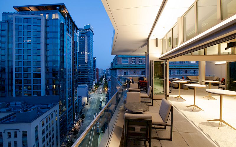 A modern cityscape at dusk with high-rise buildings and a spacious balcony featuring outdoor seating and tables, overlooking a busy street.