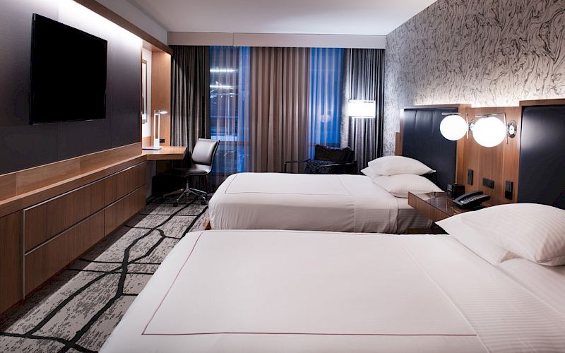A modern hotel room features two neatly made beds, a large TV, a work desk with a chair, and stylish decor with a patterned accent wall and floor lamps.