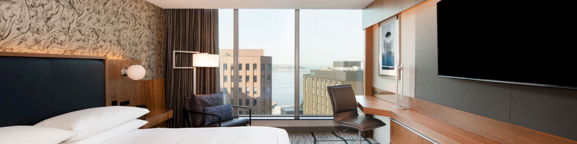 A modern hotel room featuring a large bed, a wall-mounted TV, a desk with a chair, a patterned carpet, and a window offering a city view.