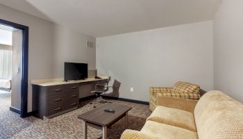 A hotel room with a desk, a chair, a TV, a coffee table, a yellow sofa, and an armchair. The room is simply furnished with neutral tones.