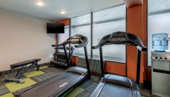 A small gym with two treadmills, a bench, dumbbells, a wall-mounted TV, and a water dispenser.