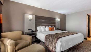 A modern hotel room featuring a cozy bed with white linens, a patterned throw, cushioned armchair and ottoman, nightstands, and soft lighting.