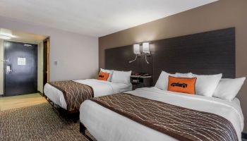 The image shows a hotel room with two neatly made double beds, a nightstand between them, and modern decor with a sleek design.