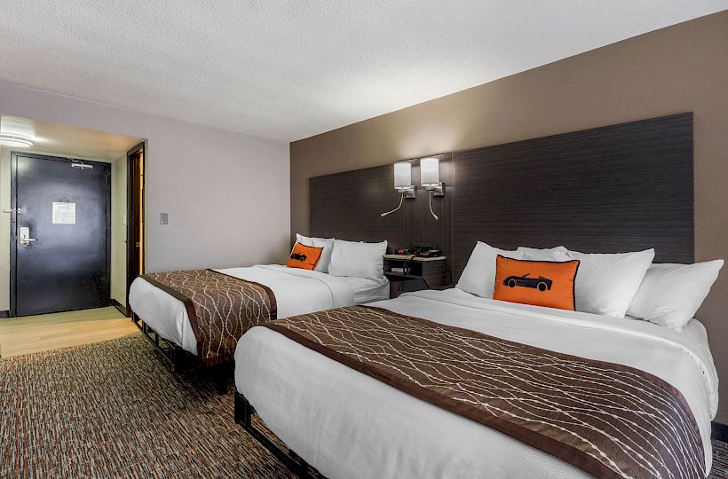 The image shows a hotel room with two neatly made double beds, a nightstand between them, and modern decor with a sleek design.