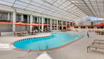 The image shows a spacious indoor pool area with multiple lounge chairs, tables with umbrellas, and a translucent ceiling letting in natural light.