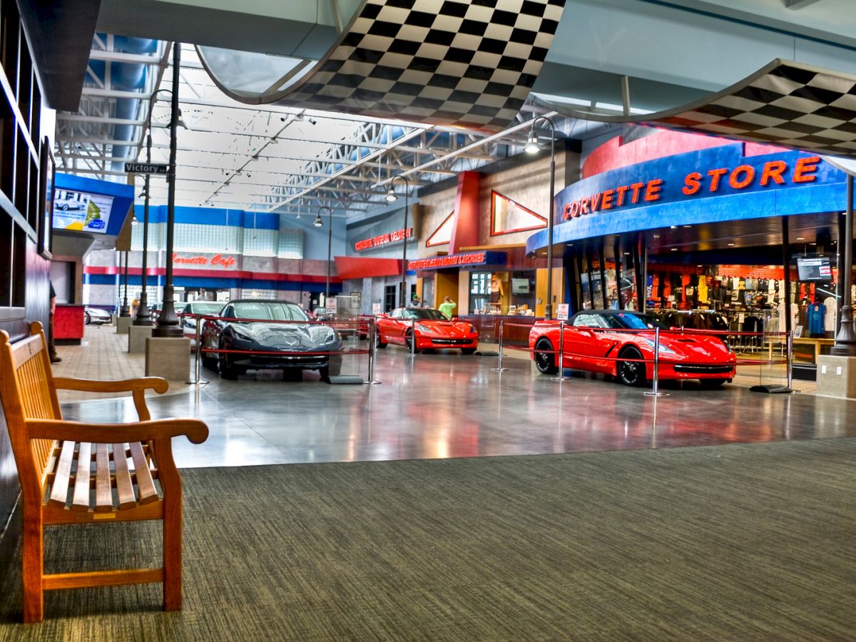 The image shows a car showroom with red sports cars, a Corvette Store, and a checkered flag ceiling. A wooden bench is in the foreground.