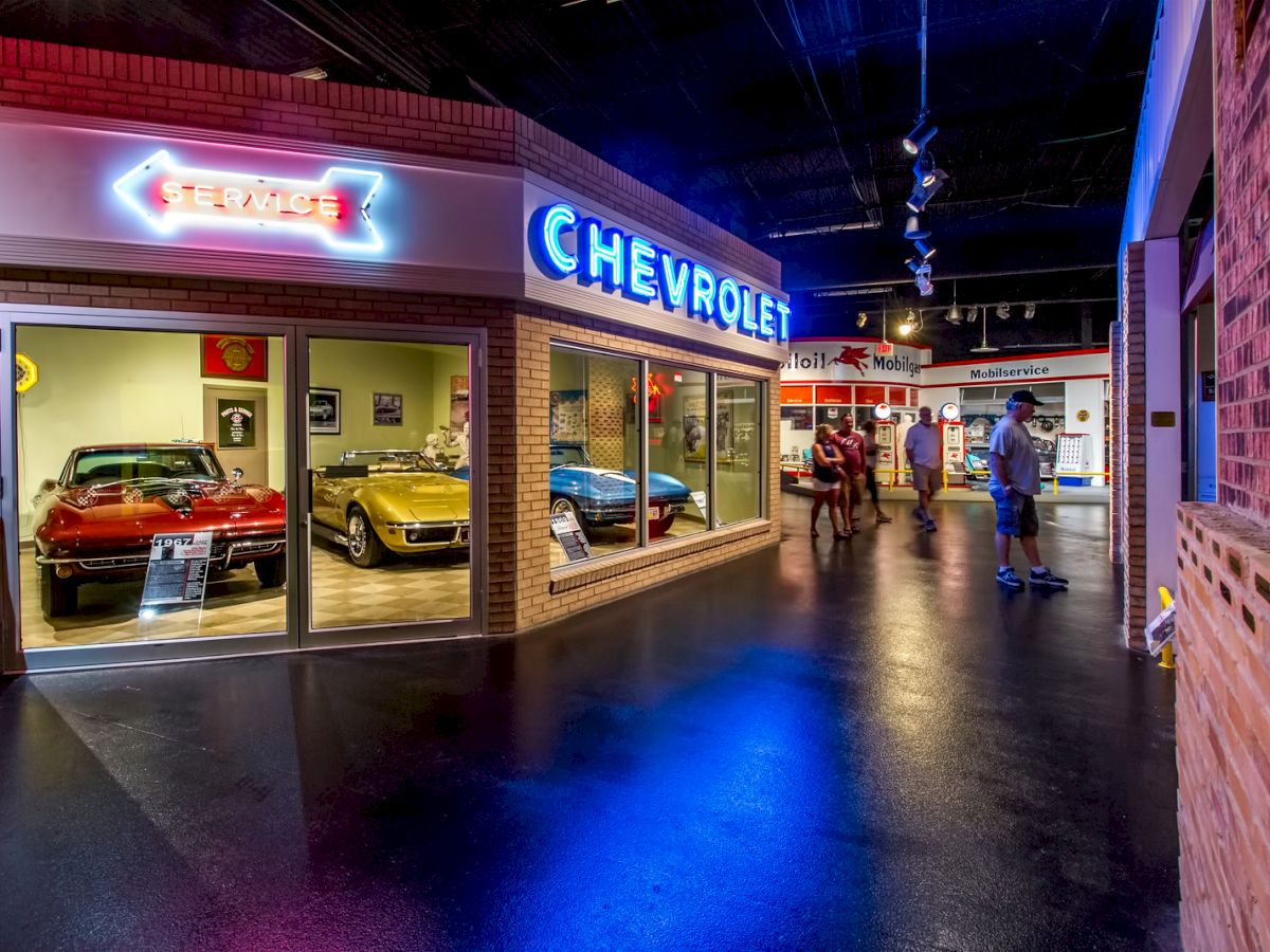 The image shows a vintage car display, focusing on Chevrolet, with neon signs. Two classic cars are in a showroom, and visitors are observing exhibits.