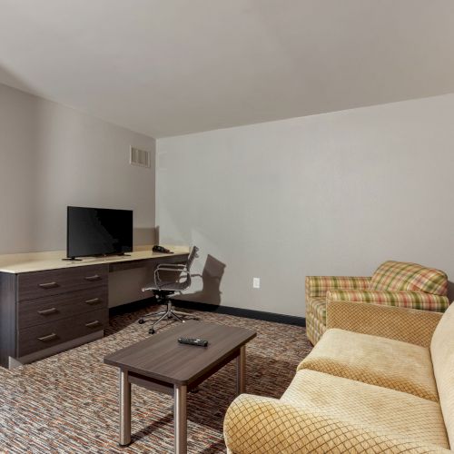 A living room with a beige sofa, plaid armchair, coffee table, desk, office chair, and a flat-screen TV on a dresser, leading into another room.