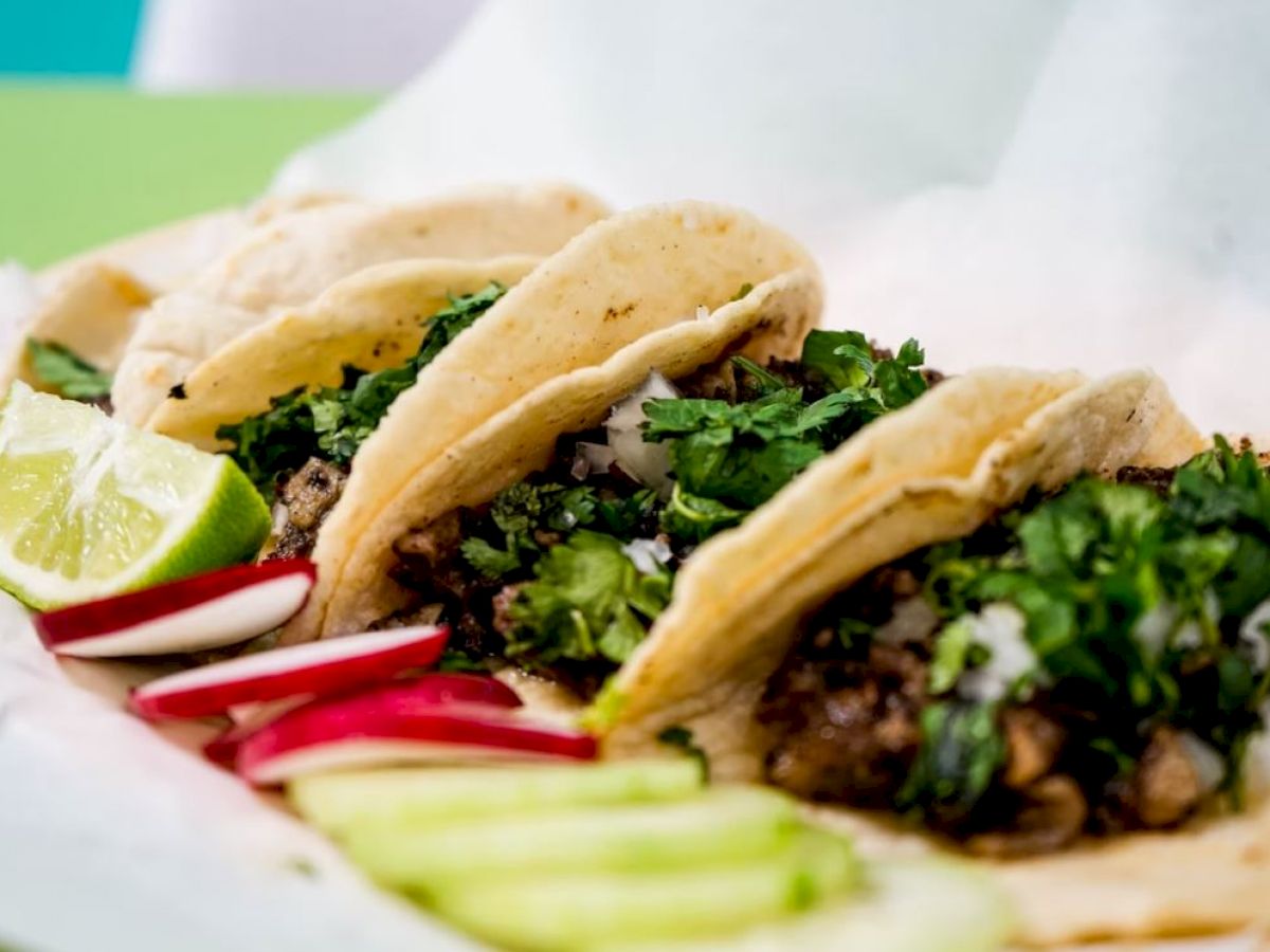 Three tacos filled with meat and topped with cilantro and onions, served with lime wedges, radish slices, and cucumber slices.