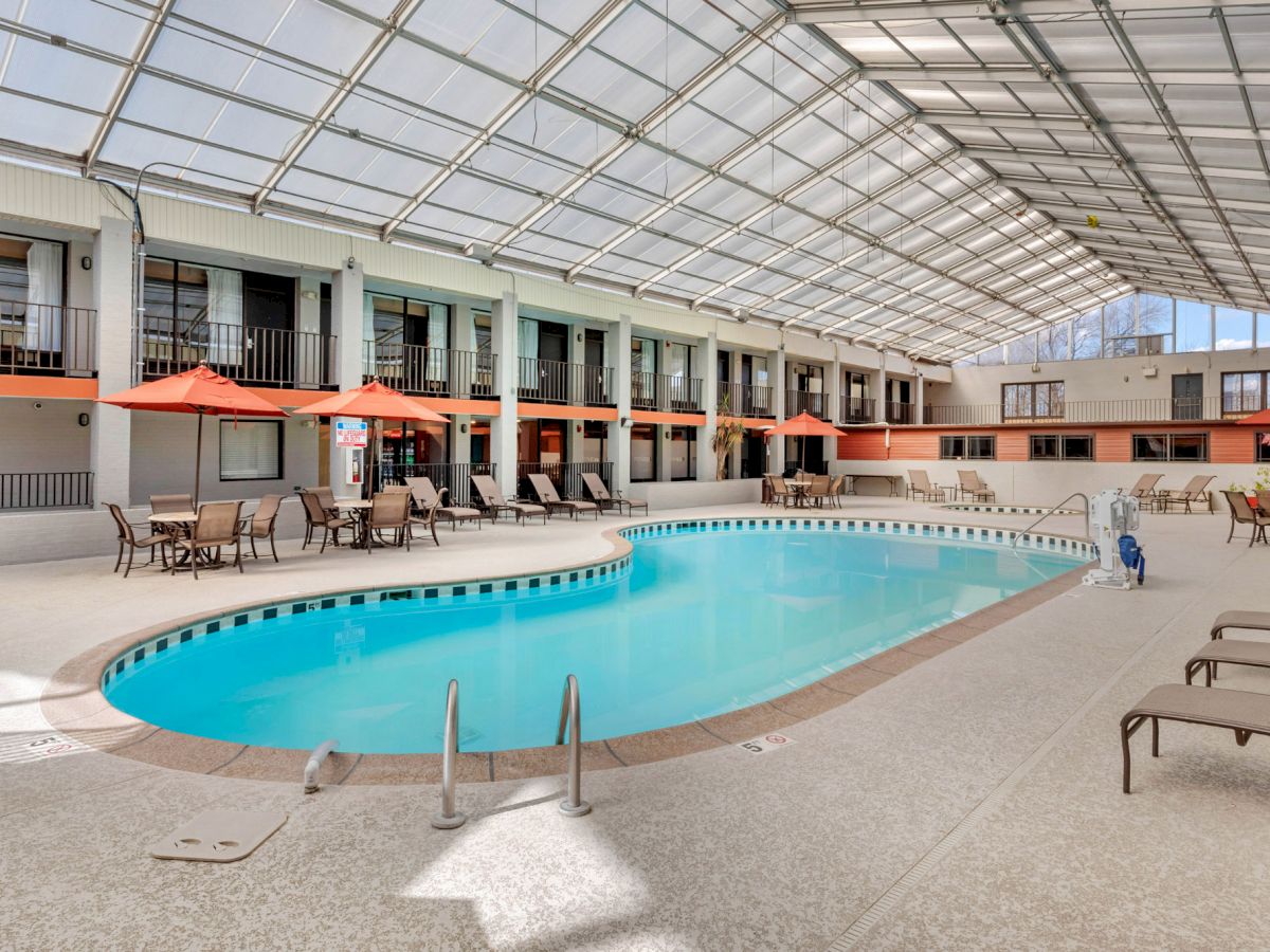 An indoor pool area with a clear roof, surrounded by lounge chairs and tables with orange umbrellas. The area appears clean and spacious.